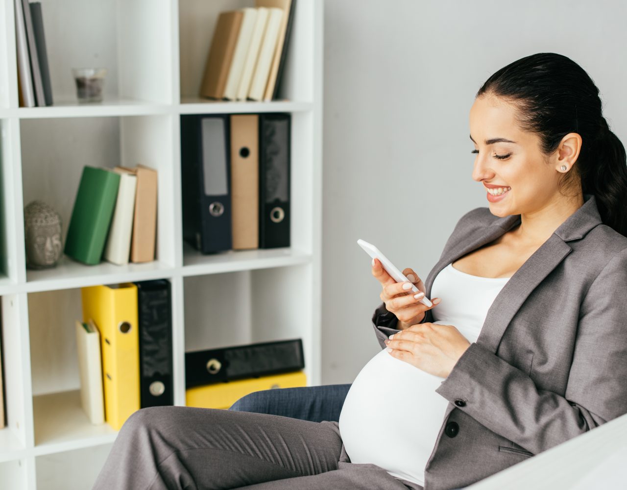 pregnant woman sitting in office chair, smiling and looking at smartphone