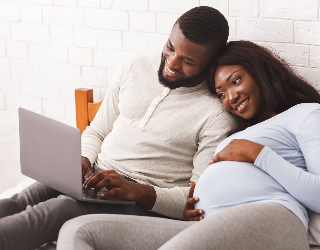 Lovely pregnant afro couple using laptop while resting at home, laying on bed, woman leaning on man shoulders, copy space